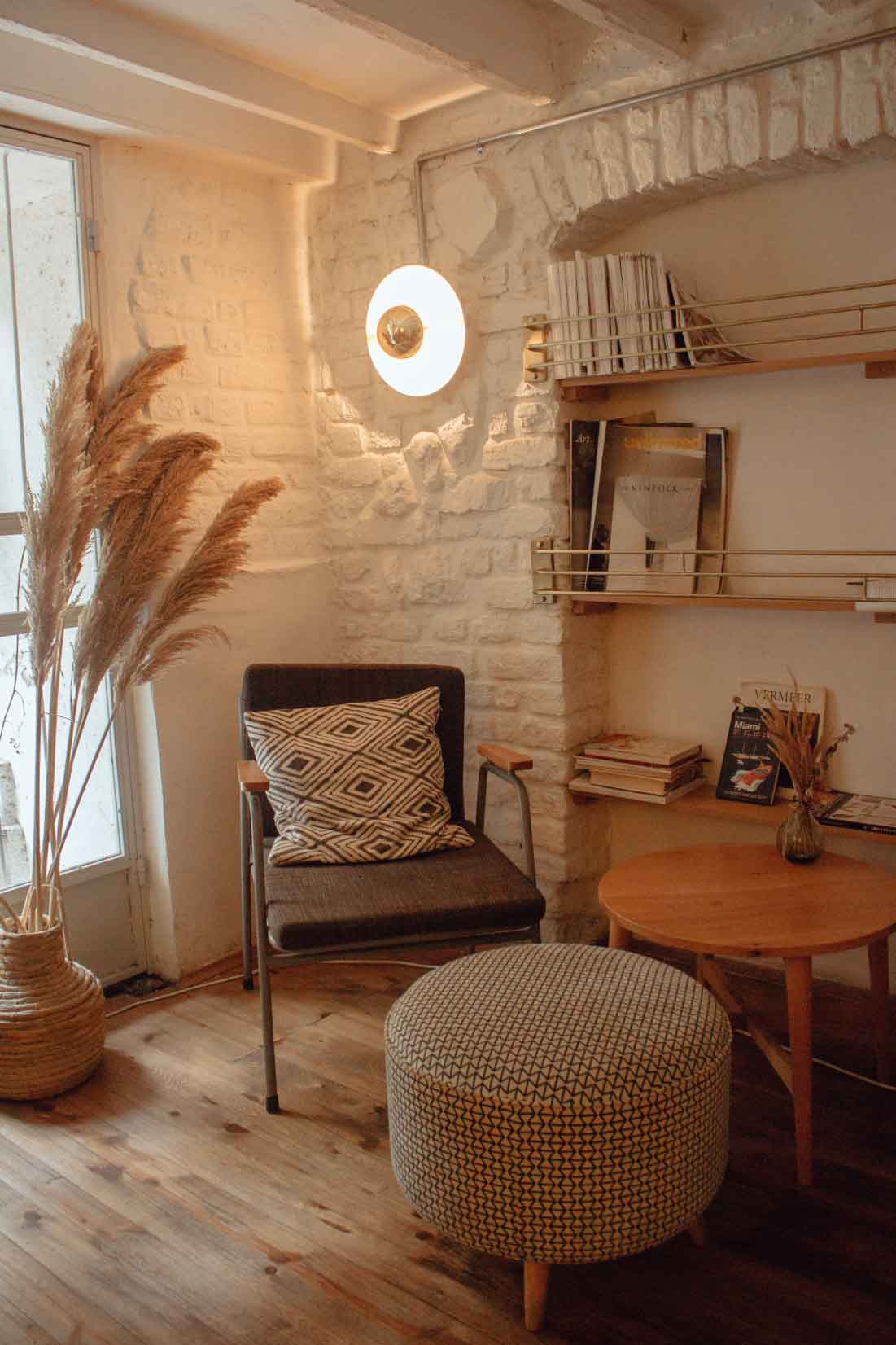 Cozy reading nook featuring a comfortable chair with a patterned cushion, a round ottoman, a tall decorative plant, and a wall-mounted light, all set against a rustic brick wall with shelves of magazines.