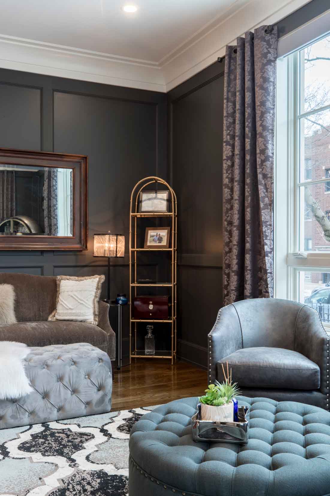 Cozy living room featuring a tufted gray ottoman, a plush sofa with decorative pillows, a stylish gray armchair, and a gold shelving unit displaying personal items, set against dark walls and large windows with elegant curtains.