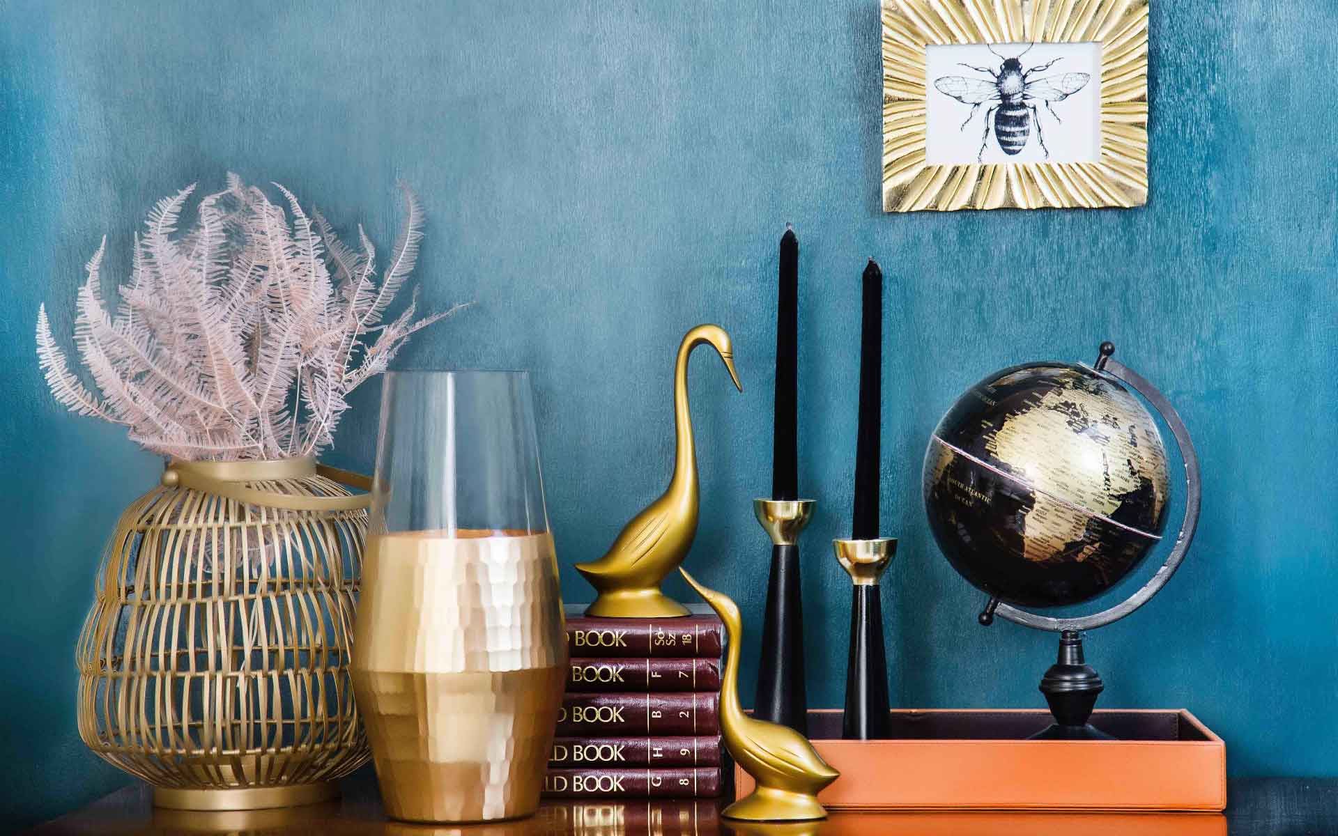 Decorative home accents including a golden vase with pink foliage, a sleek glass vase, gold and black candlesticks, a globe, and a framed bee illustration against a teal background, emphasizing home decor and energy conservation themes.