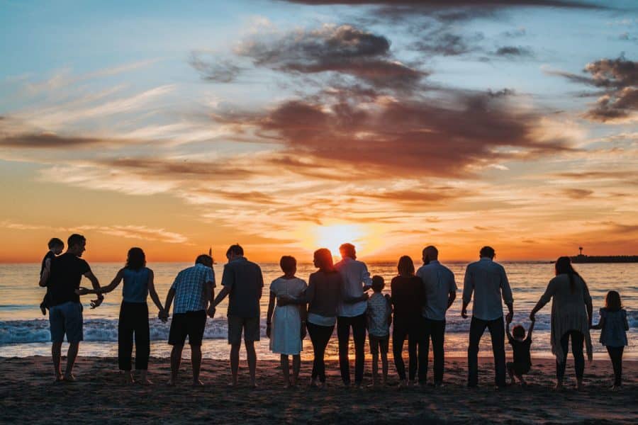 Family standing together on the beach at sunset, holding hands, with the ocean and colorful sky in the background, reflecting themes of unity and togetherness.